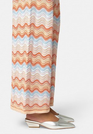 Piernas bajas con una falda de patrón ondulado y multicolor en tonos pastel y zapatos plateados bajos de tacón con punta afilada y sin cordones sobre un fondo blanco.