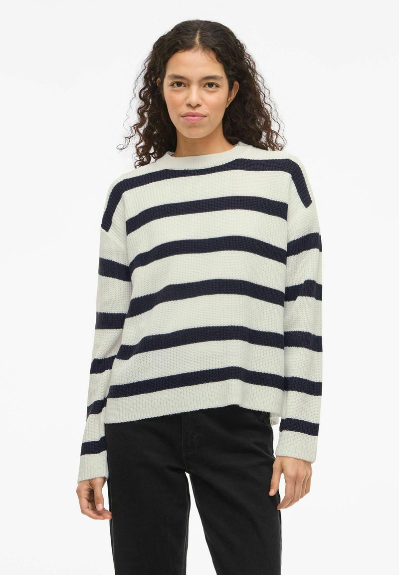 Pull rayé crème et marine, avec une texture côtelée, un col rond et une coupe décontractée, associé à un pantalon noir.