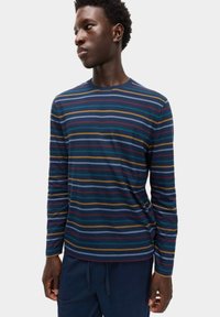 Giovane uomo che indossa una maglietta a maniche lunghe blu navy con sottili righe orizzontali in giallo, teal, bordeaux e azzurro, in piedi contro uno sfondo bianco.