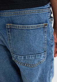 Jeans in denim blu con una grande tasca posteriore, cuciture visibili e una texture leggermente consumata. La vita è scura e aderente.