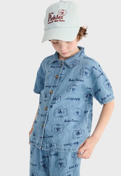 Enfant portant une chemise en denim bleu clair et un short avec des motifs de visages de chiens dessinés, ensemble assorti, et une casquette blanche avec le texte "Pickles" et une illustration de chien.