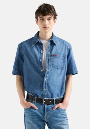 Giovane uomo che indossa una camicia di jeans blu a maniche corte sbottonata sopra una t-shirt bianca, jeans blu e una cintura nera, in piedi con le mani nelle tasche.