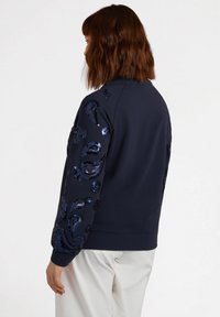 Sudadera azul marina con patrones de paisley de lentejuelas en las mangas, escote redondeado y puños de canalé. Tejido suave con un corte relajado.