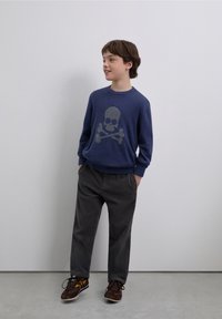 Suéter azul marino con diseño de calavera y huesos cruzados, pantalones de pana grises y zapatillas marrones con detalles amarillos. Modelo en pose casual.