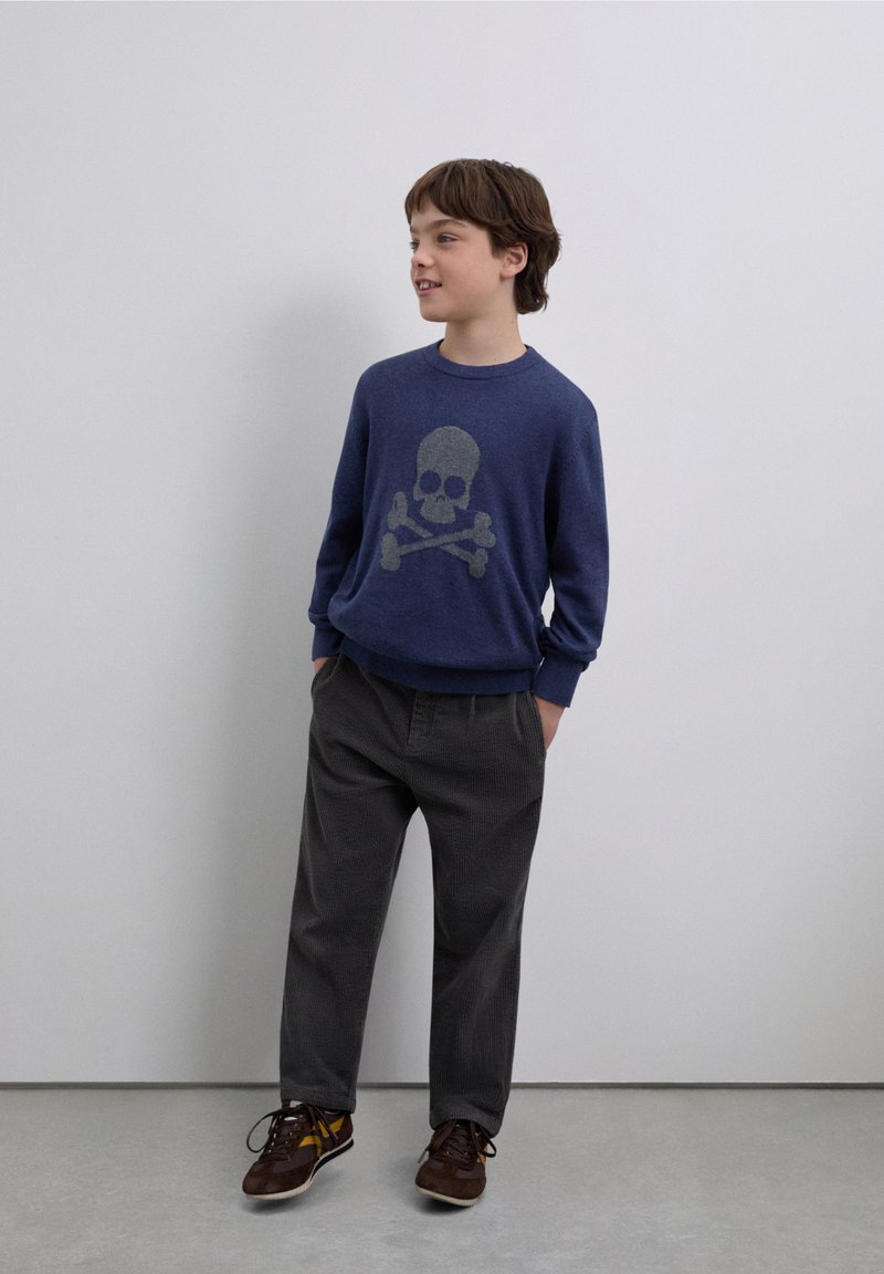 Suéter azul marino con diseño de calavera y huesos cruzados, pantalones de pana grises y zapatillas marrones con detalles amarillos. Modelo en pose casual.