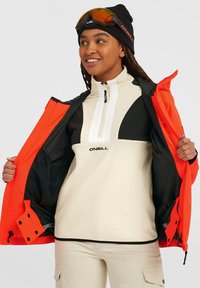 Vrouwen O'Neill fleece pullover in crème en zwart, met een witte rits, gecombineerd met een oranje jas. Heeft een gestructureerde stof en ritsen.