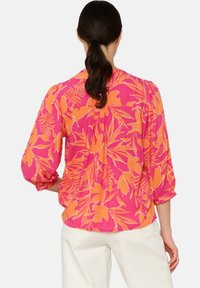 Bluse mit floralem Muster auf pinkem Hintergrund, die große orangefarbene Blätter zeigt. Lange Ärmel mit Raffung, leichtes Material und lockere Passform.