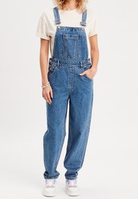 Salopette en denim à coupe décontractée, couleur bleu clair, bretelles réglables, poche avant et boutons sur les côtés. Associée à des baskets blanches.