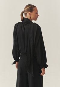 Čierna hodvábna blúzka s dlhými nadýchanými rukávmi a viazaním na chrbte. Hladká textúra, zúžený výstrih, v kombinácii s čiernymi nohavicami s vysokým pásom.