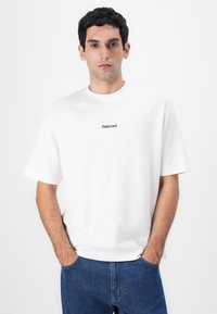 Hvit bomulls t-skjorte med korte ermer, med en liten svart "Selected" logo brodert på brystet, sammen med blå denimjeans.