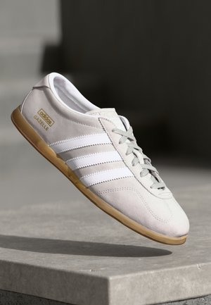 White Adidas Gazelle sneaker with gum sole and gray laces, mid-air above a concrete surface with a dark blurred background.