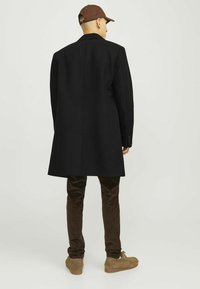 Abrigo de lana negro con un diseño estructurado, visto desde atrás. Combinado con pantalones de pana marrones y calzado beige. Gorra marrón sencilla.