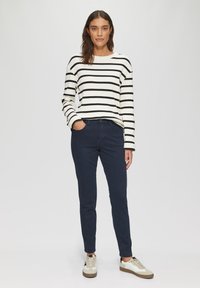 Pull rayé à manches longues en blanc et noir, associé à un pantalon marine et des baskets beiges. Le pull a une texture côtelée.