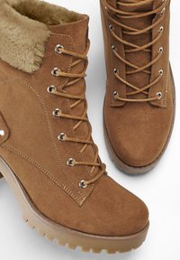 Stivali ankle in suede marrone con fodera in pelliccia sintetica, dotati di occhielli in metallo e design con lacci, suola in gomma rinforzata con texture antiscivolo.