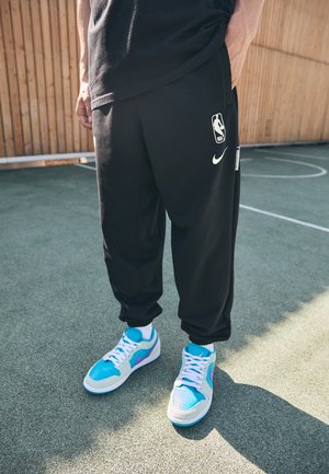 Osoba nosząca czarne spodnie dresowe NBA oraz niebiesko-białe trampki stoi na zewnętrznym boisku do koszykówki na tle drewnianego płotu.