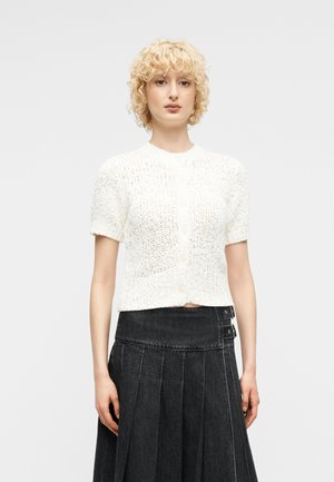 Ξανθιά γυναίκα με σγουρά μαλλιά, που φοράει ένα άσπρο πλεκτό ζακέτα με κοντά μανίκια και μια μαύρη πλισέ φούστα ψηλόμεση με λεπτομέρειες αγκράφας.