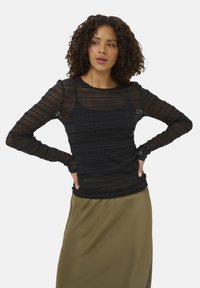 Haut noir à manches longues avec un design en dentelle texturée superposée, comportant des sections côtelées et transparentes, associé à une jupe verte olive.