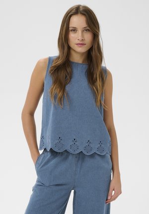 Kobieta o długich brązowych włosach, ubrana w niebieski top bez rękawów z falbankowym kwiatowym wycięciem na dole oraz dopasowane niebieskie spodnie, stojąca z rękami w kieszeniach.