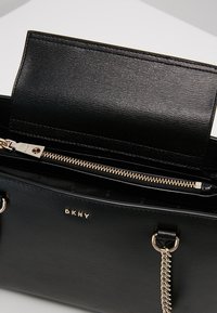 Sac à main noir DKNY en cuir texturé, avec une fermeture éclair dorée, une bandoulière en chaîne et un compartiment intérieur visible sous le rabat ouvert.
