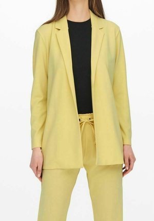 Donna che indossa un blazer giallo chiaro e pantaloni coordinati con una maglietta nera sotto, in piedi su sfondo bianco.