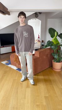 Pitkähihaine ruskea paita, jossa on logo, vaaleat farkut ja hopeiset lenkkarit. Huoneessa on ruskea corduroypussikangas-sohva ja suuri vihreä kasvi.