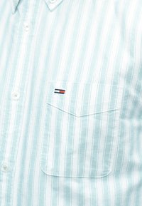 Ανοιχτό μπλε και λευκό πουκάμισο με κάθετες ρίγες, κουμπωτό, με τσέπη στο στήθος που διαθέτει ένα μικρό κόκκινο, λευκό και μπλε λογότυπο.