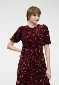 Φούστα από μπορντώ παγιέτα με κοντά, φουσκωτά μανίκια. Υφή που λαμπυρίζει λόγω των παγιέτων. Κλασική στρογγυλή λαιμόκοψη, μήκους μέχρι το γόνατο.