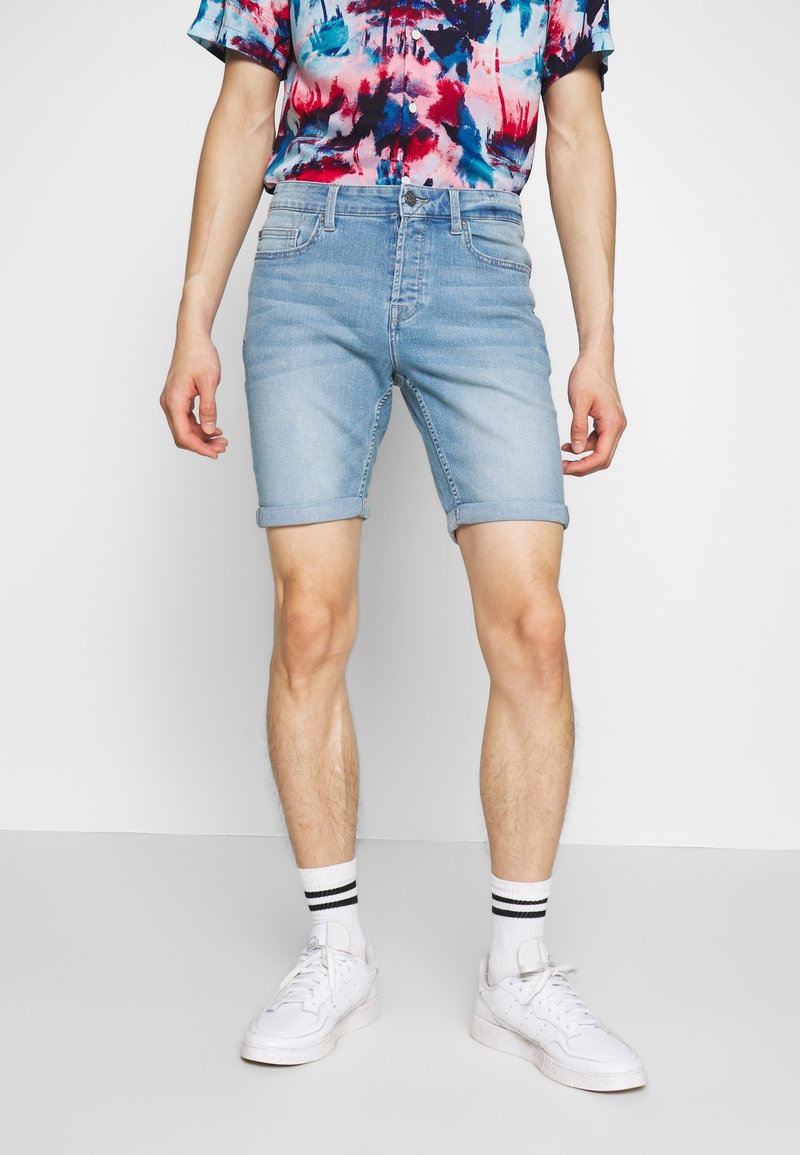 Personne portant un short en denim bleu clair, des baskets blanches, des chaussettes blanches à rayures noires, et une chemise à manches courtes colorée, debout devant un fond uni.