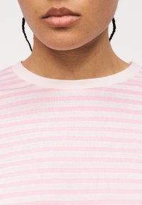 Κοντινό πλάνο του κάτω μέρους του προσώπου και του λαιμού ενός ατόμου που φοράει ριγέ ροζ και άσπρη μπλούζα με στρογγυλή λαιμόκοψη και εμφανείς πλεγμένες τούφες μαλλιών.