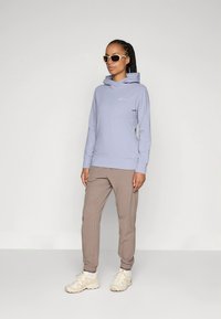 Sweatshirt com capuz roxo claro com bolso canguru e logótipo, combinado com calças beje afuniladas e ténis de cor clara.