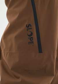Braune Skihosen mit glatter Textur, ausgestattet mit einem marineblauen Reißverschluss und einem auffälligen schwarzen Logo an der Seite. Verstärkter Nahtdetail sichtbar.