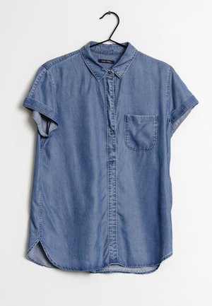 Niebieska koszula dżinsowa z krótkim rękawem, z przednimi guzikami, jedną kieszenią na piersi i kołnierzykiem, wisząca na czarnym wieszaku na białym tle.