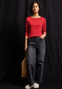 Rotes Langarmoberteil, hochtaillierte dunkelgraue Jeans und hellfarbige Sneaker. Hält eine beigefarbene Tragetasche und steht vor einem dunklen Hintergrund.