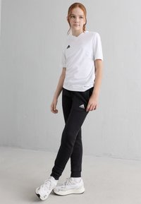 Fiatal személy áll, fehér Adidas pólót, fekete Adidas melegítőt és fehér tornacipőt visel egy sima szürke fal előtt.