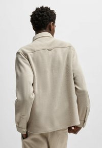 Veste en velours côtelé beige avec un design structuré, un détail de empiècement au dos et des poignets à boutons, présentant une surface texturée et une coupe décontractée.