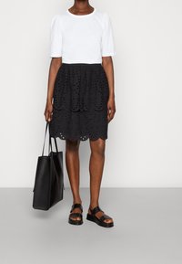 Haut blanc à manches courtes associé à une jupe noire en eyelet avec un ourlet festonné, portant un sac cabas noir et des sandales noires.