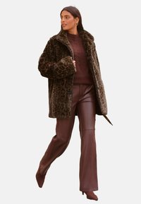 Kabát z umelého kožúška s leopardím vzorom a širokými klopami, v kombinácii s pleteným burgundským svetrom a hnedými koženými zvonovými nohavicami. Hnedé platformové topánky.