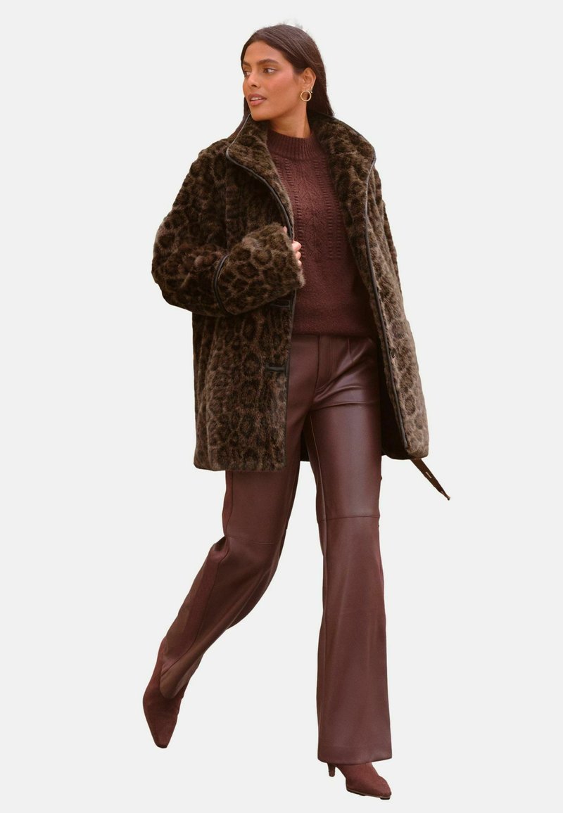 Kabát z umelého kožúška s leopardím vzorom a širokými klopami, v kombinácii s pleteným burgundským svetrom a hnedými koženými zvonovými nohavicami. Hnedé platformové topánky.