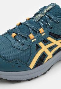 Plava i žuta Asics tenisica za trčanje s mrežastim materijalom, ojačanim prstom, uzorkastim vezicama i strukturiranom sivom potplatom, pod kutom.