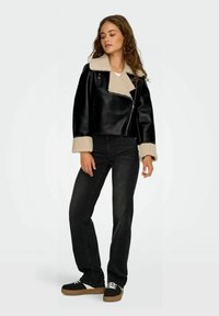 Giacca corta in pelle nera con collo e polsini in pelliccia sintetica beige. Il modello indossa jeans neri e sneakers nere con suole chiare.