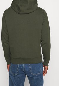 Felpa verde oliva con cappuccio, polsini e bordo a coste. Realizzata in tessuto morbido e texturizzato, indossata con jeans denim azzurri.