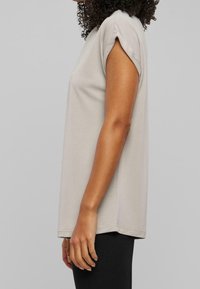 Lysegrå t-shirt med korte, rullede ærmer, afslappet pasform og blød tekstur. Stylret med sorte leggings, set fra siden.