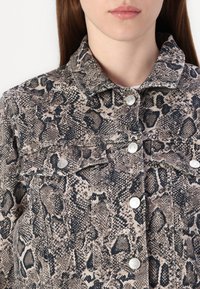 Turtleneck denimjakke med slangeskinnmønster i beige og svart, med sølvfargede trykknapper og brystlommer.