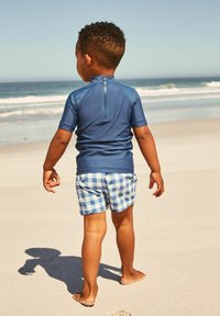 Blå kortærmet rash guard med lynlås på ryggen, parret med lyseblå ternede badebukser med grønne accenter, båret på en sandstrand.