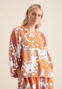 Blusa ampia bianca con grandi motivi floreali arancioni e blu, caratterizzata da un collo a V e maniche a sbuffo, realizzata in un tessuto leggero.