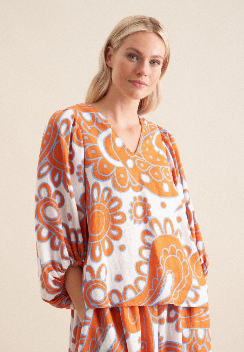 Blusa ampia bianca con grandi motivi floreali arancioni e blu, caratterizzata da un collo a V e maniche a sbuffo, realizzata in un tessuto leggero.