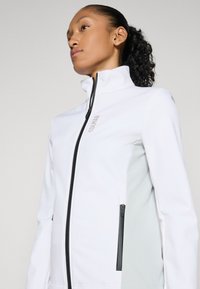Weiße Softshelljacke mit hohem Kragen, schwarzem Reißverschluss und grauen seitlichen Akzenten; verfügt über ein Logo auf der Brust und Reißverschlusstaschen.
