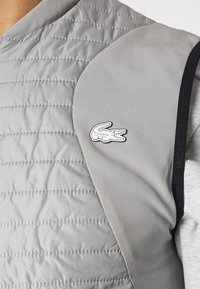 Grå vadderad väst med quiltad textur, med rund hals och en Lacoste krokodil-logotyp på vänster axel. Slät texturfärdig.