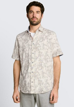 Mann trägt beige kurzärmeliges Hemd mit abstraktem Blumenmuster, kombiniert mit hellgrauer Hose, steht vor schlichtem Hintergrund.
