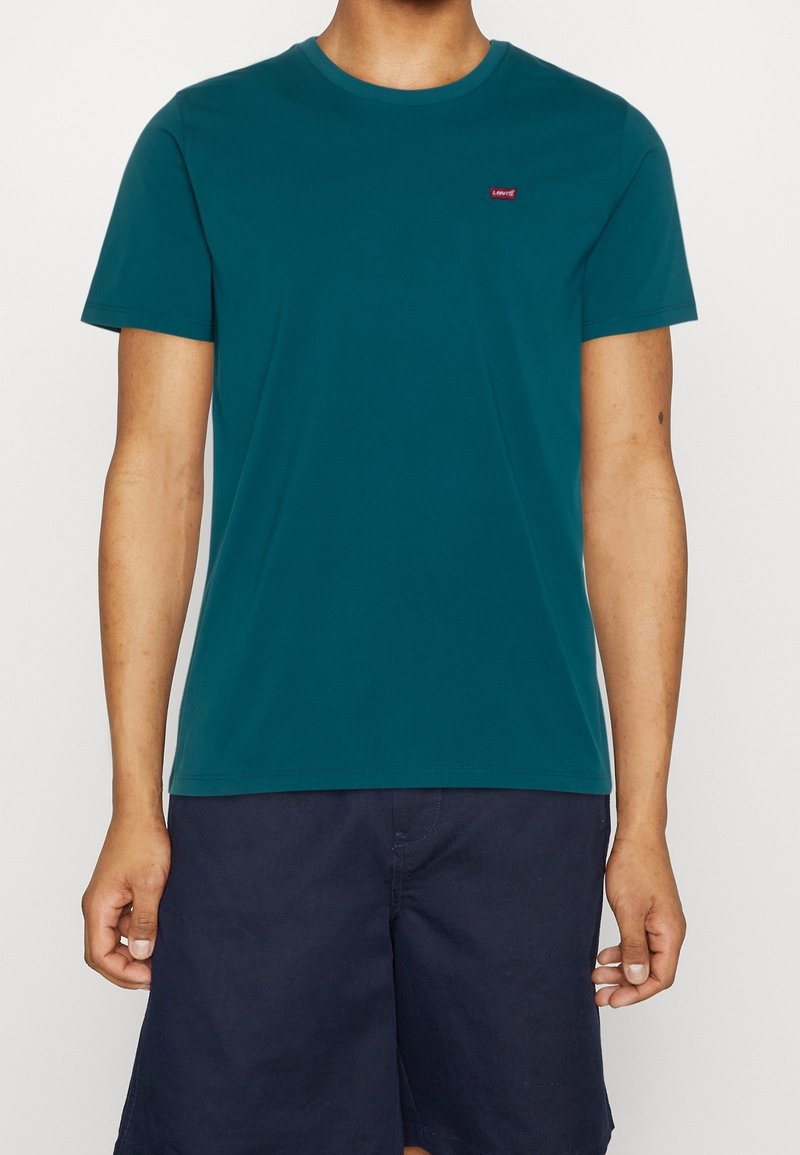 Personne portant un t-shirt uni manches courtes couleur sarcelle avec un petit logo rouge Levi's sur la poitrine et un short bleu marine.